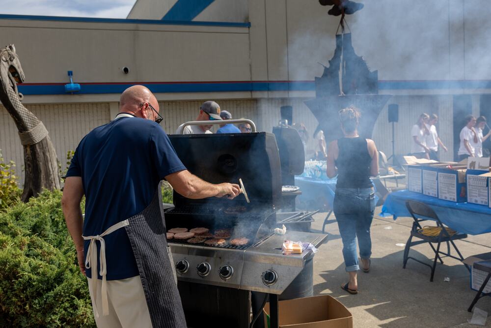 A volunteer cooking on the grill, smoke coming off the lid, with tables of food set up behind.