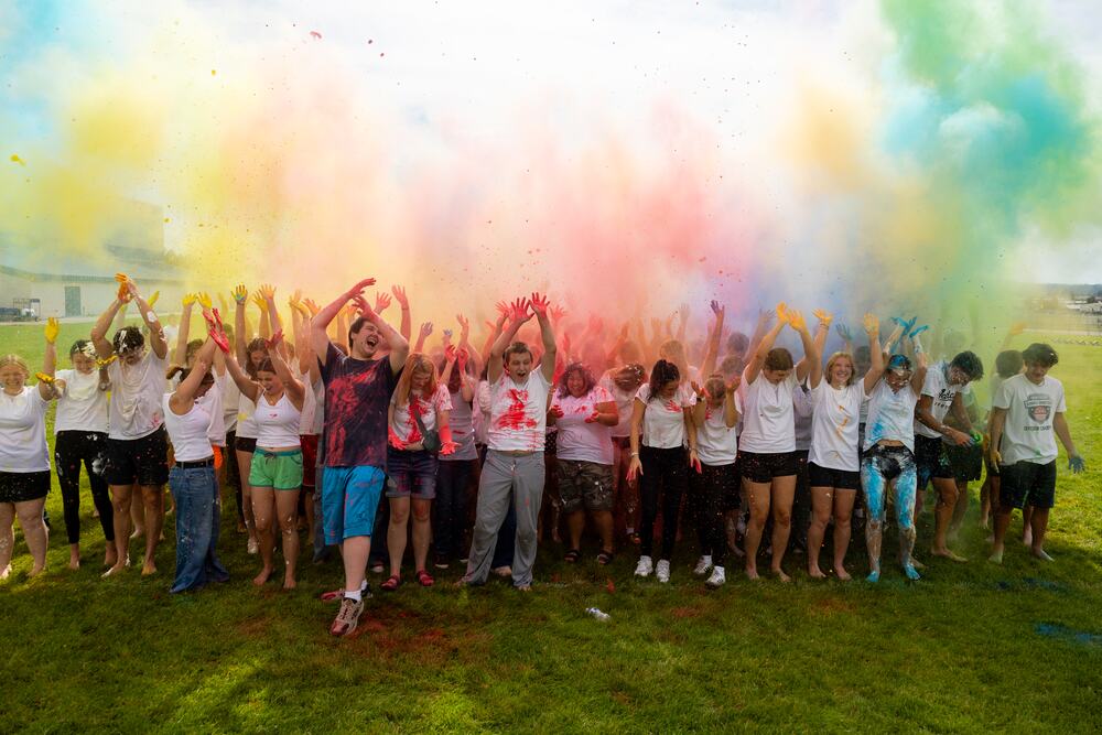 The senior class standing together with their arms raised under a wall of red, yellow, blue and green color powder.